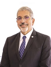A man with grey hair wears glasses, a black suit jacket, white button down, and blue and white tie