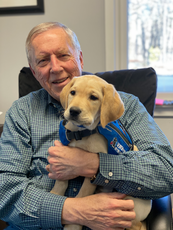 A man wearing a plaid button down holds a golden retriever puppy waring a blue service vest