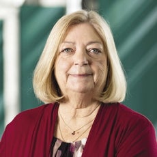 A woman with chin length blonde hair wears a red sweater and black, white, and purple patterned blouse.