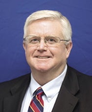 A headshot of a man with white hair wearing glasses, a black suit jacket, white button down, and red, yellow, black, and blue striped tie