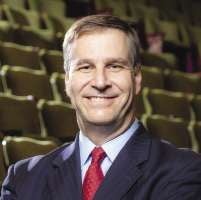 A man with short light brown hair wears a black suit jacket, white button down, and red tie with theater chairs behind him.