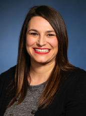A headshot of a woman with long brown hair wearing pink lipstick, a black cardigan, and grey top