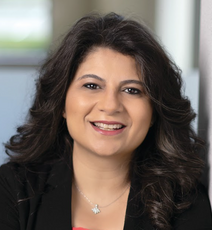 A woman with curled dark hair wears a black suit jacket and a silver necklace.