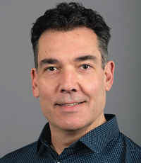 A man with dark brown hair wears a dark blue patterned button down.