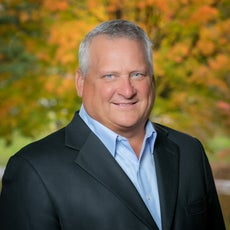 A man with grey and white hair wears a black suit jacket and a light blue button down.