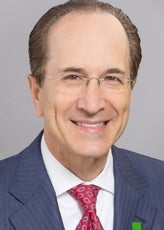 A man with light brown hair wears rimless glasses, a pinstriped dark blue suit jacket, a white button down, and a red paisley tie.