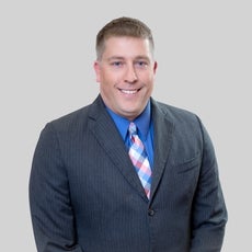 A man with short blonde hair wears a grey pinstriped suit jacket, a blue button down, and a blue, pink, and white tie.