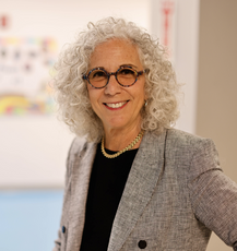 A woman with curly white hair wears circle glasses, a black and white tweed suit jacket, a black top, and a gold chain necklace.