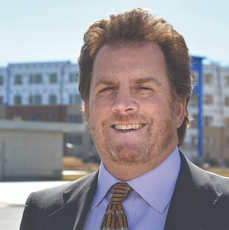 A man with red hair and a red beard wears a black suit jacket, a purple and white patterned button down and an orange, black, and white patterned tie while standing in front of buildings under construction.