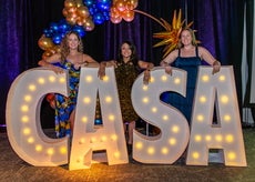 Three women stand behind large lit-up letters spelling C-A-S-A