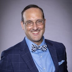 A man with short grey hair wears wire-rimmed glasses, a blue plaid suit jacket, a light blue button down, and a blue, white, and black plaid bow tie.