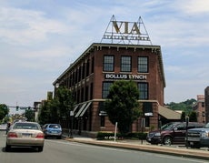 A brick building with a sign reading "Bollus Lynch" on it sits between a busy street on the left and a parking lot on the right.