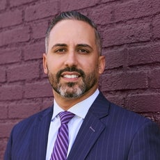 A man with short salt and pepper hair and beard wears a dark blue pinstriped suit jacket, a light blue button down, and a purple and pink striped tie while standing in front of a brick wall.