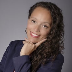A woman with long curly brown hair wears a dark blue blazer and stands with her hand under her chin and her pointer finger on her cheek.