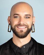 A person with a beard, large square earrings, and a black shirt smiles at the camera.