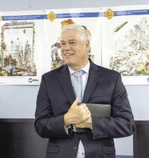 A man with white hair wears a black suit jacket, a white button down, and a grey tie while holding a book to his chest.
