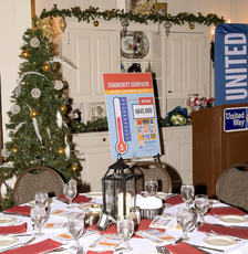 A round table with glasses and red napkins sits in front of a Christmas tree and a sign with an image of a thermostat partially filled in to represent money raised during a campaign