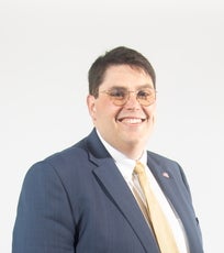 A man with glasses wears a blue suit with a white shirt and yellow tie.