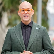 A bald man with orange glasses wears a black button down and a green tweed suit jacket