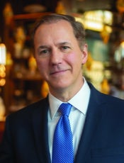 Headshot of a man with light brown/grey hair wearing a dark blue suit jacket, a white button down, and a blue patterned tie