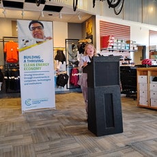 Woman speaks at podium in a bike shop with biking clothes and accessories behind her.
