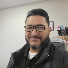 A man with black hair and a black beard wears black glasses and a black jacket and black polo with a white undershirt.