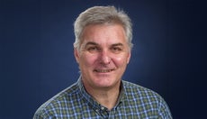 A man with grey hair wears a blue, green, and white plaid button down.
