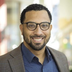 A man with short dark hair wears black glasses, a grey blazer, and a blue button down.