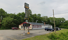 A small liquor store surrounded by a parking lot with a large sign out front