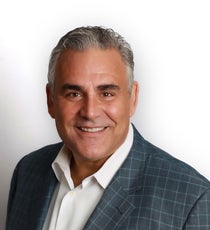 A man with short white and grey hair wears a blue plaid suit jacket and a white button down.