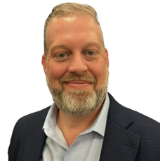 A man with grey hair and a beard wears a black suit jacket and a light blue button down.
