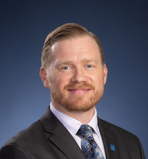A man with short hair wears a black suit jacket, a light purple button down, and a blue patterned tie.