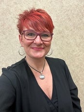A woman with short red hair wears red glasses and a black top.