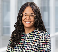 A woman with long, curly brown hair wears glasses and a tweed jacket.