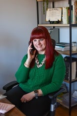 A woman with red hear in a green sweater sits at a desk while holding a cell phone to her ear and smiling