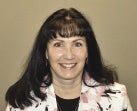 A woman with long dark hair and bangs wears a pink and white floral blazer and a black top.