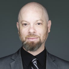 Headshot of a bald man with a brown and grey beard wearing a dark grey suit jacket, black button down, and black and grey tie