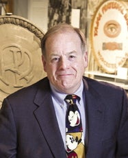 A man wears a dark blue suit jacket, a light blue button down, and a Mickey Mouse tie.