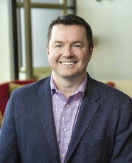 A man wears a dark blue suit jacket and purple striped button down.