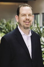 A man with dark hair, a beard, a white shirt, and a blue jacket