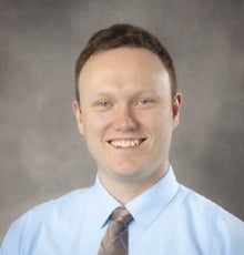A man wearing a blue shirt and brown tie smiles for the camera.