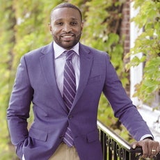 A man wearing a blue suit jacket, white and blue striped button down and tie stands outside with his hand on a railing