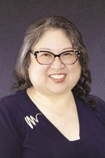 A woman with shoulder length hair wears glasses and a purple blouse.