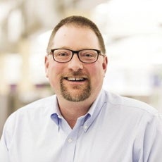 A headshot of a man with black glasses and a goatee wearing a light blue button up