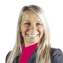 A woman with long, straight blonde hair wears a grey blazer and a pink turtleneck.