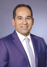 A headshot of a man with a dark blue suit jacket, white button down, and light blue tie