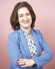 A woman with shoulder-length brown hair wears a light blue blazer, standing with her arms folded.