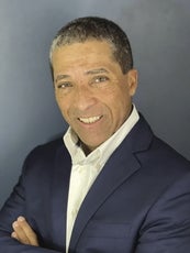 A man wears a white button down and a blue blazer, standing with his arms crossed.