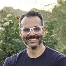 A man with a dark beard and white glasses wears a blue T-shirt.