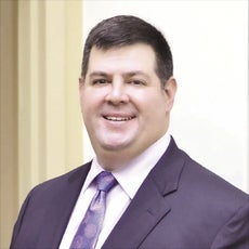 A man with short dark hair wears a black suit jacket, white button down, and blue patterned tie.